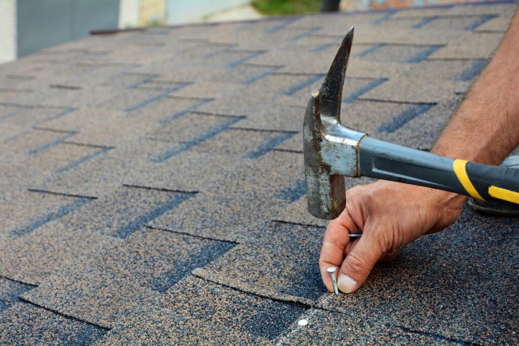 Roofer is hammering a Nail in the Roof Shingles. How Much Does A Shingle Roof Cost?