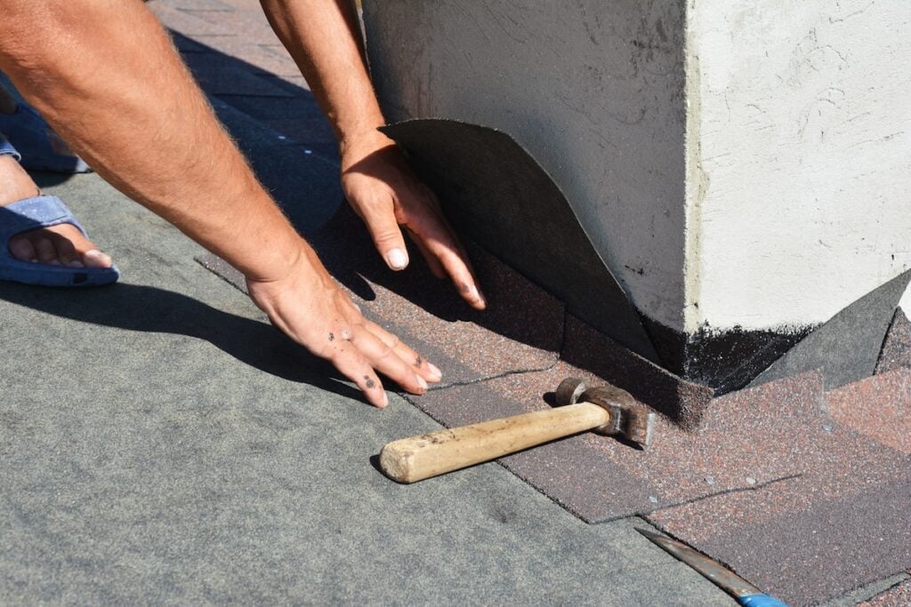 Asphalt shingles roofing construction. Roofer repair and laying asphalt shingles with vapor barrier in chimney problem area. Chimney waterproofing on house rooftop.