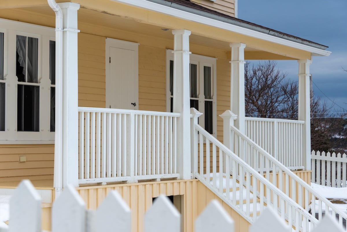 A roofed porch of a country style yellow and white house. There are multiple double hung windows and a wooden shutter door. The overhanging roof has a number of pillars and fence enclosing the deck.