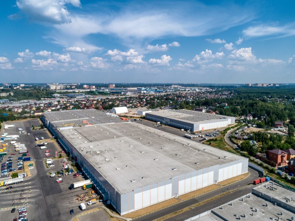 Huge warehouse building with flat roof and cars on parking site under blue sky with fluffy clouds near city in summer bird eye view commercial roof cleaning