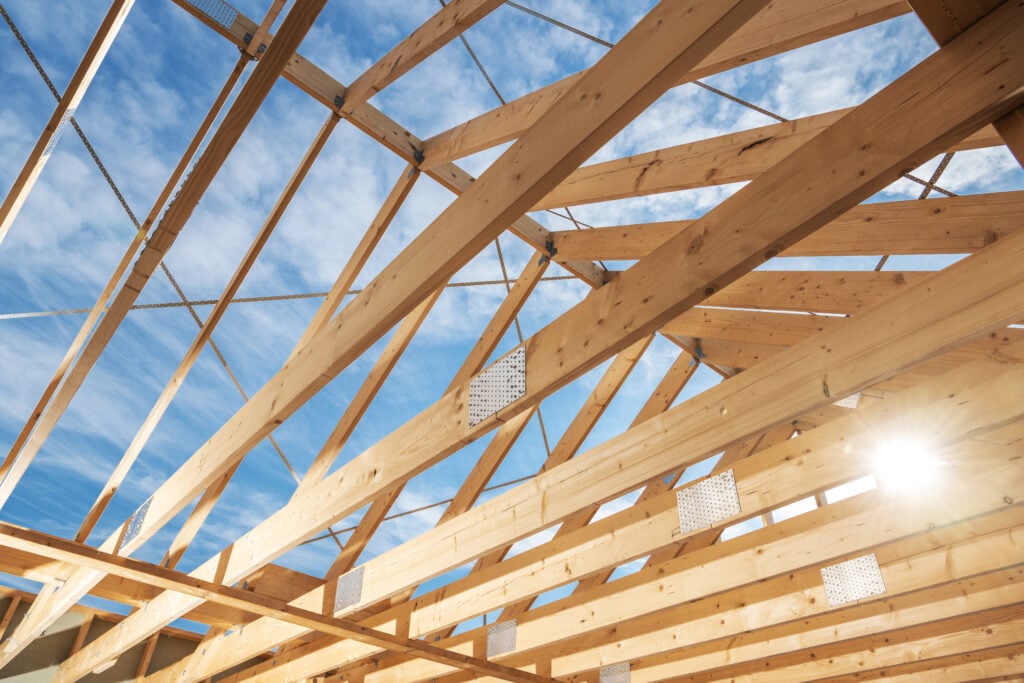 The framework of a wooden roof is visible against a bright blue sky, showcasing beams and trusses in the construction process.