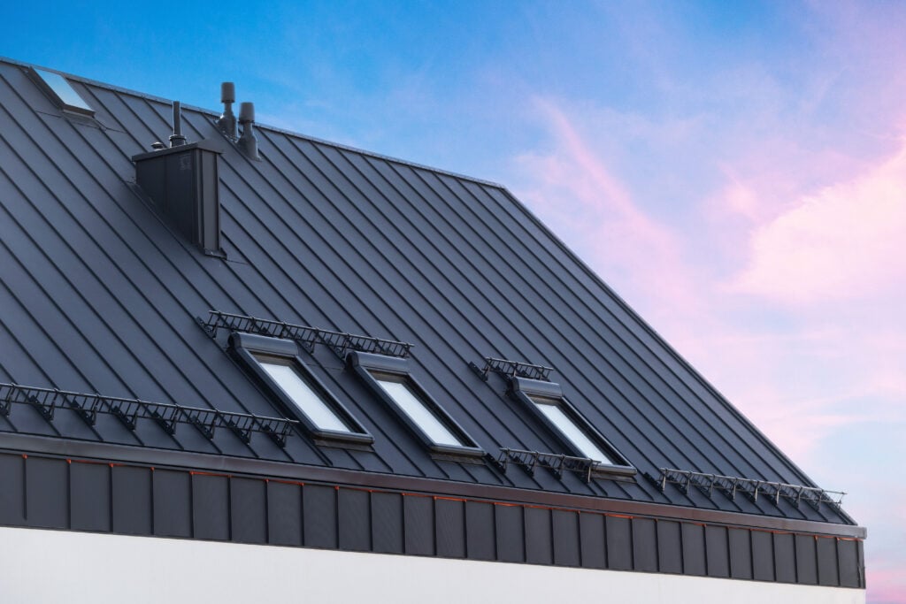 mansard roof replacement cost House with metal galvanized coating, chimney pipe, mansard windows and snow holding cornice on roof slope against sunset sky. Element of modern residential building with zinc rooftop sheets