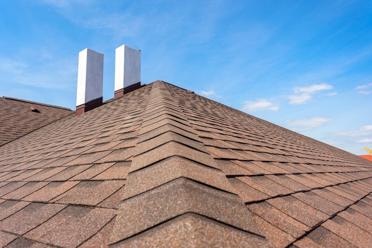 Photo of new roof with asphalt tile and two white chimney on new home under construction, against blue sky
