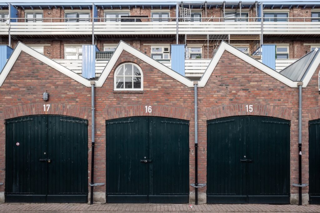 Outdoor street view in front of exterior garage entrance with wooden green doors, brick facade and sawtooth gable roof of European apartment or housing.