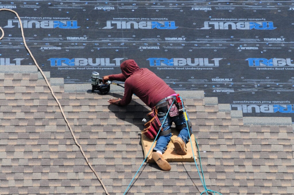 Roofer at work using a pneumatic nail gun to install shingles over a synthetic underlayment on a building’s roof
